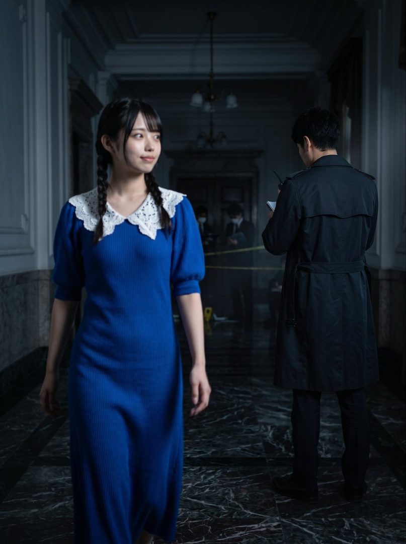 A young woman in a blue dress with a white lace collar and braided hair walks through a dimly lit hallway, with a man in a trench coat in the background.