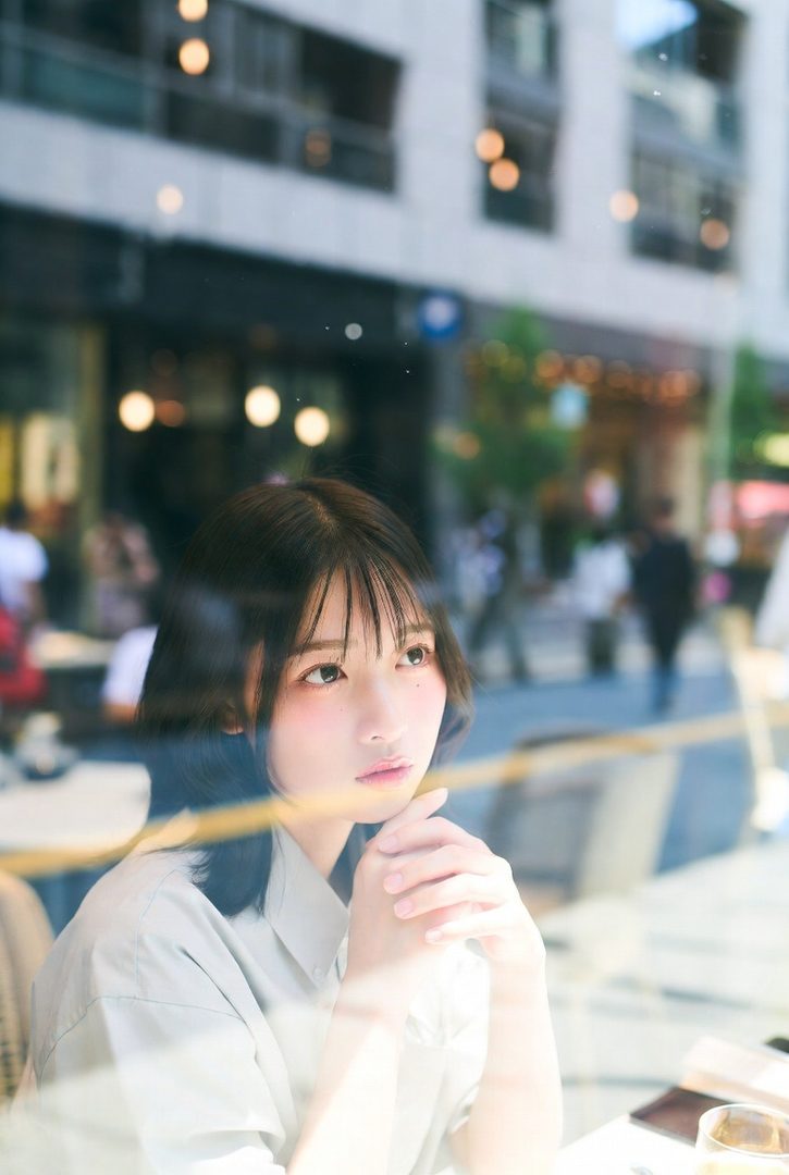 A young woman with short black hair and bangs sits in a cafe, looking out the window with a serene expression, with reflections on the glass.