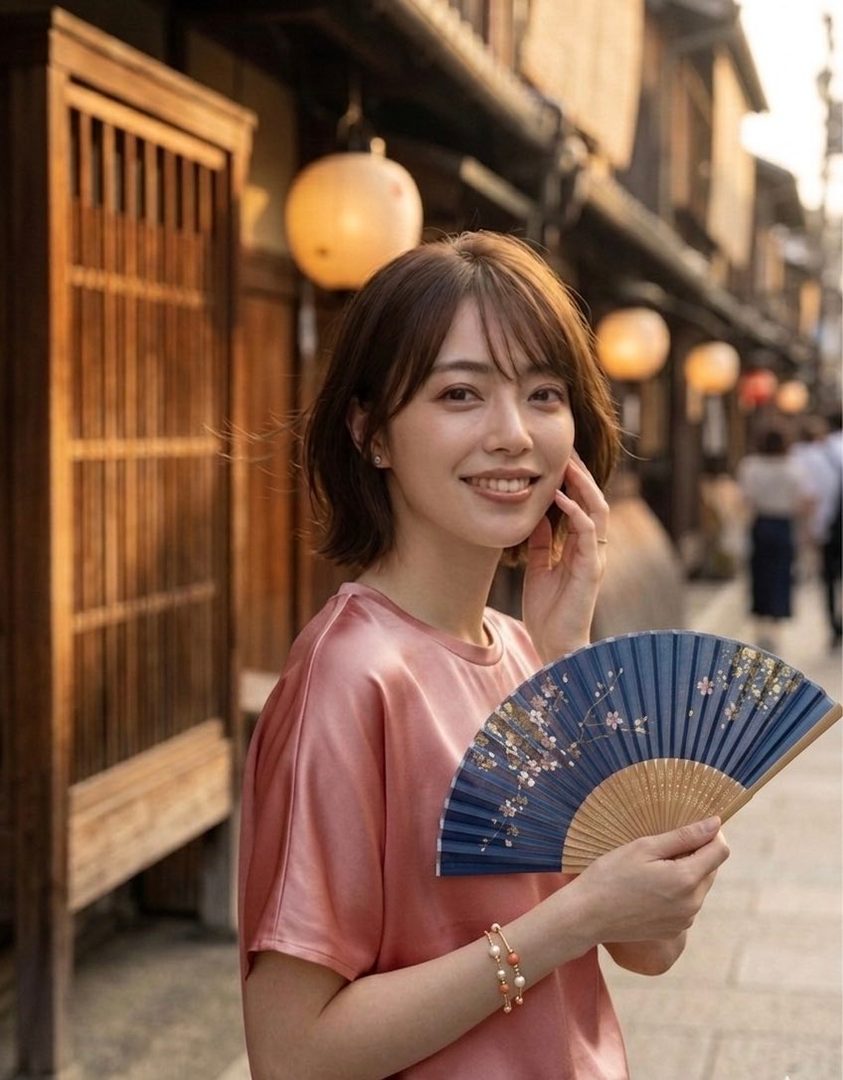A young woman with short brown hair smiles gently while holding a blue fan with cherry blossom designs on a traditional Japanese street.