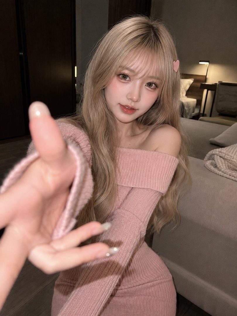 A young woman with long blonde hair and bangs, wearing a pink off-shoulder knit top, extends her hand towards the camera in an indoor setting.