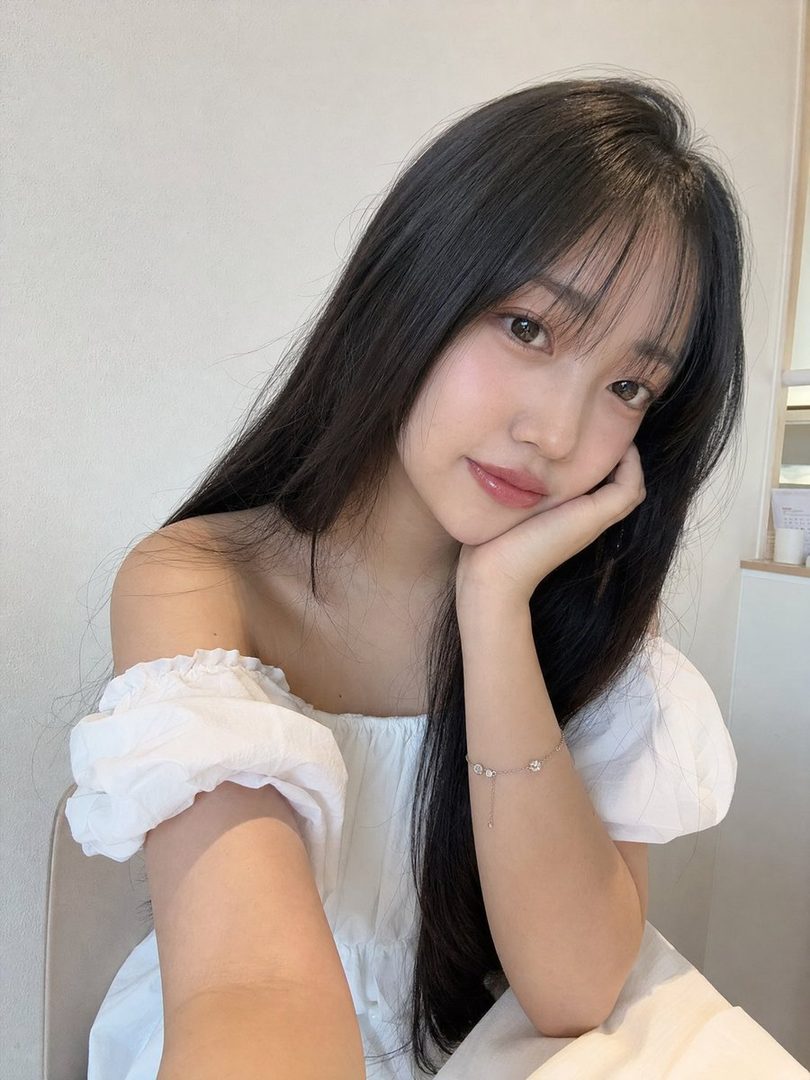 A young woman with long black hair and bangs, wearing a white off-shoulder blouse, poses for a selfie indoors.