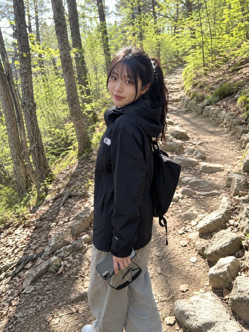 A young Japanese woman with black hair tied in a ponytail, wearing a black jacket and light gray pants, stands on a forest trail.