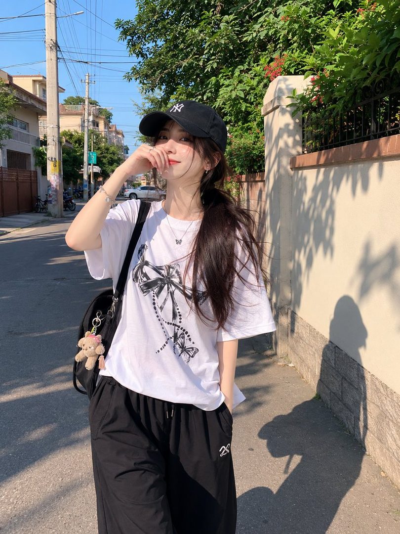 A young woman with long dark hair and a black baseball cap walks down a sunny residential street, wearing a white graphic t-shirt and black pants.