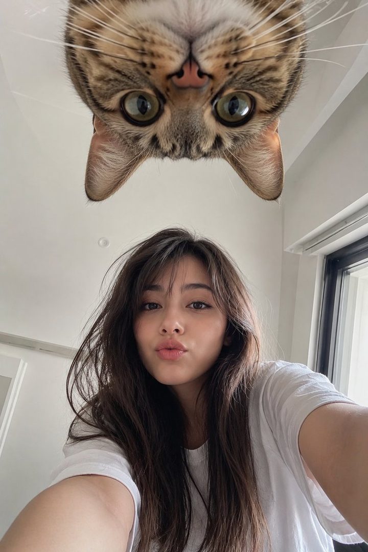 A young Japanese woman takes a selfie with a cat looking down from above, both making cute expressions.