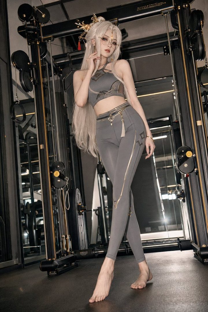 A woman with long blonde hair and glasses, dressed in a grey sports bra and leggings with gold accents, poses barefoot in a gym.