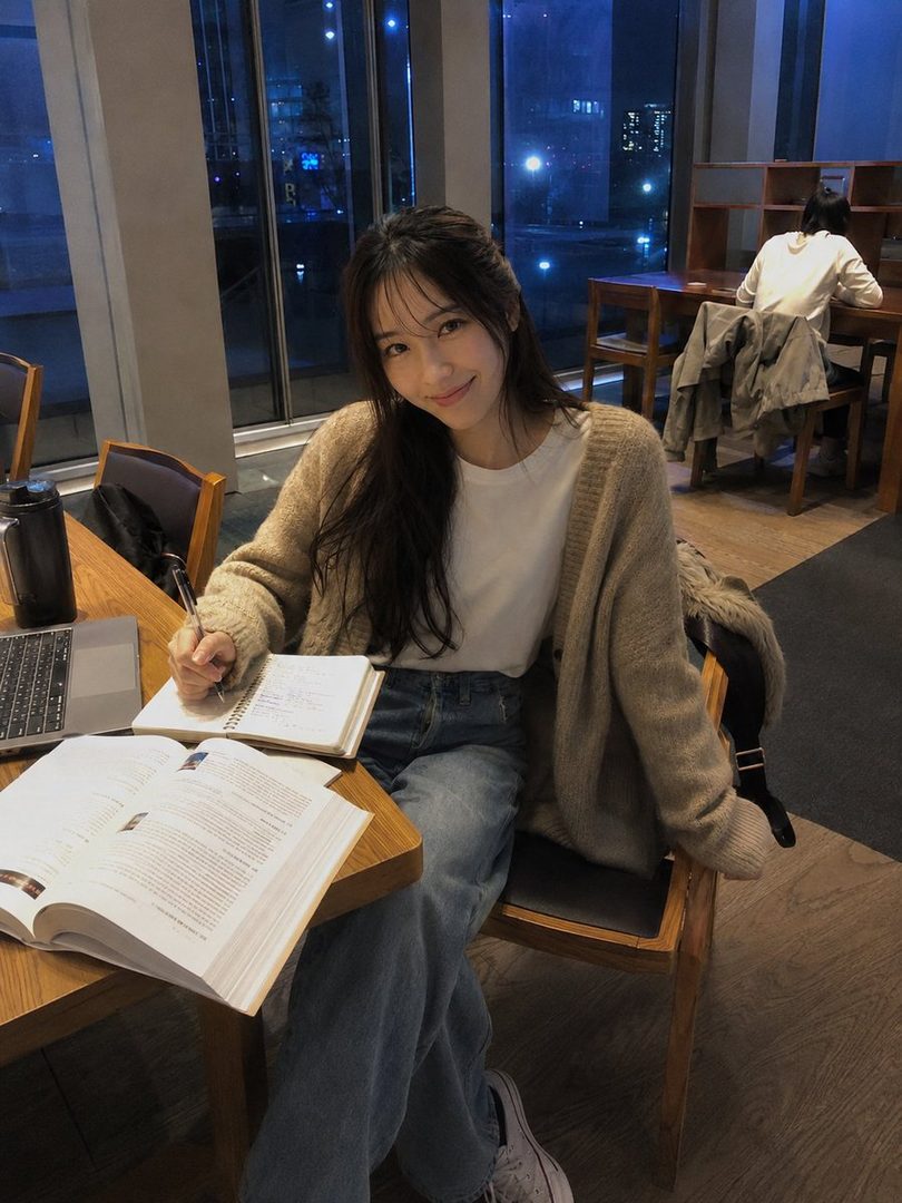 A young woman with long dark hair smiles at the camera while studying at a cafe table at night, wearing a beige cardigan and jeans.