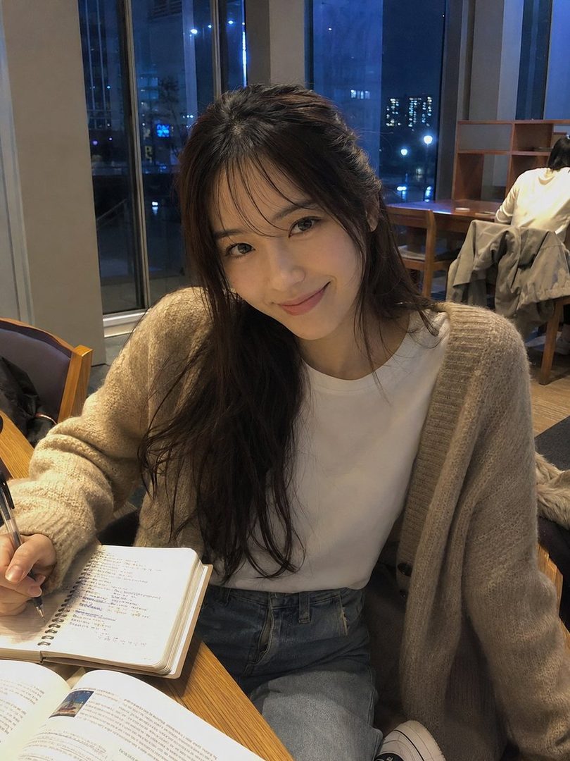 A young woman with long dark hair and bangs, wearing a beige cardigan and white t-shirt, smiles while studying in a cafe at night.