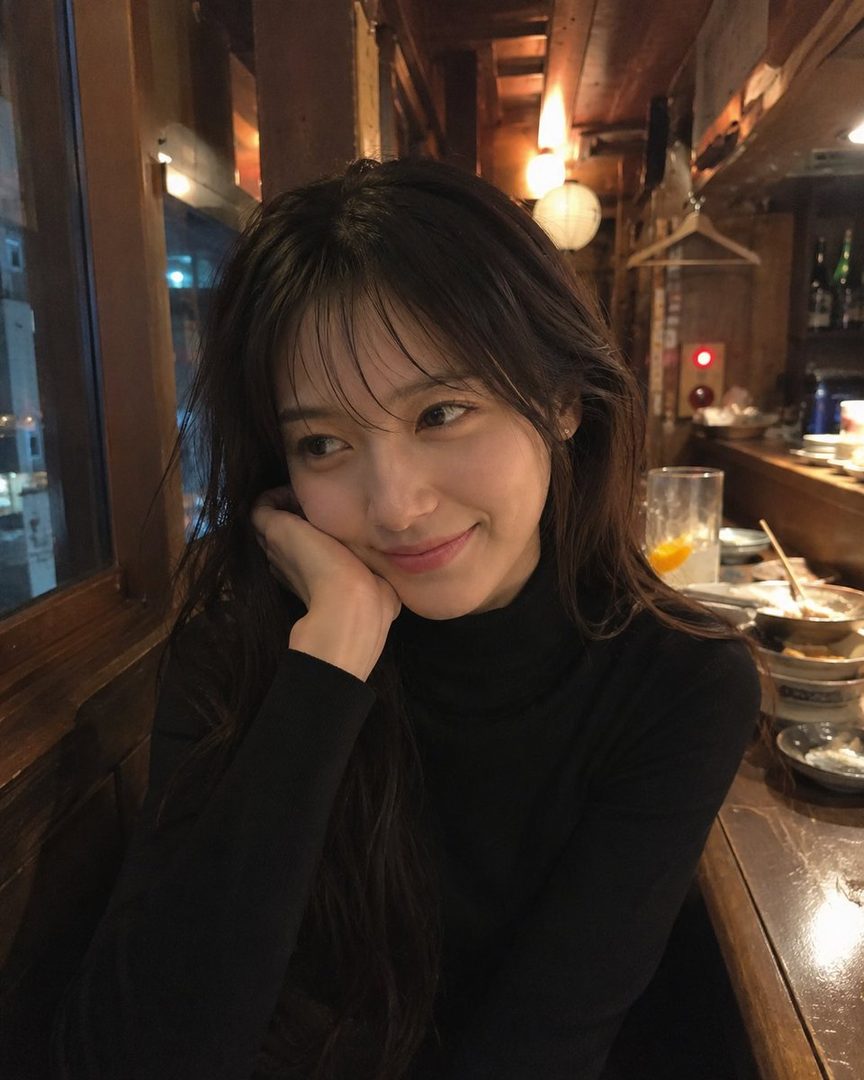 A young woman with long dark hair and a black turtleneck smiles gently while resting her chin on her hand in a dimly lit izakaya.