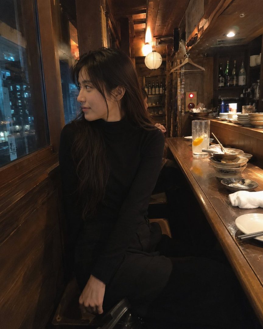 A young woman with long dark hair wearing a black turtleneck sits at a wooden bar in a dimly lit izakaya, looking out a window at the city lights.