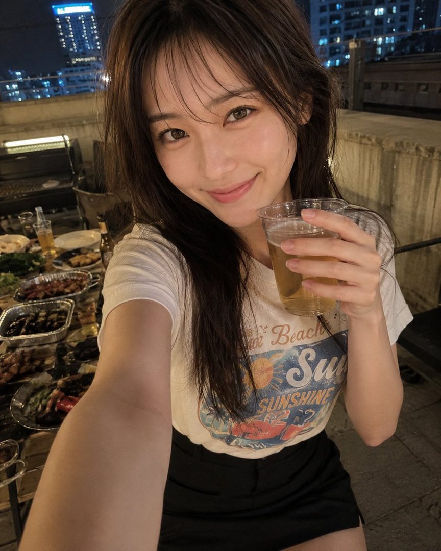 A young woman takes a selfie while holding a drink at a rooftop BBQ at night, with city lights in the background.