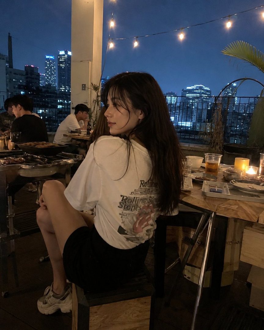 A young woman with long dark hair sits at a table on a rooftop at night, with city lights in the background.