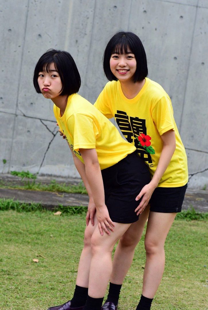 Two Japanese women in yellow t-shirts and black shorts pose playfully outdoors on a grassy area with a concrete wall in the background.