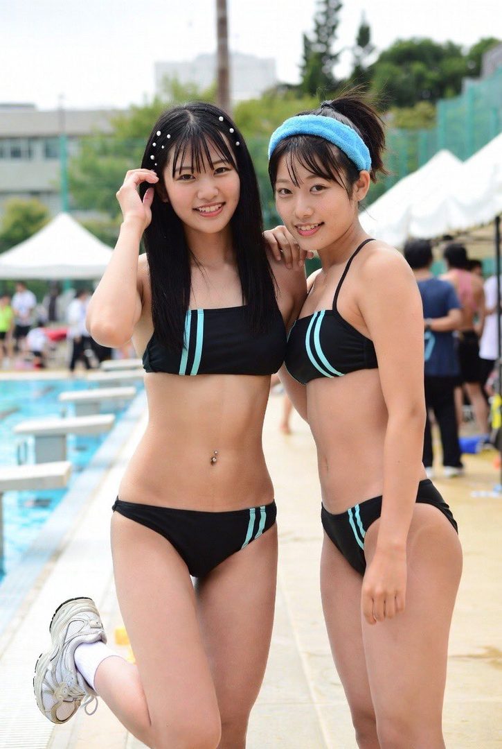 Two young Japanese women in black bikinis with blue stripes pose cheerfully by an outdoor swimming pool on a sunny day.