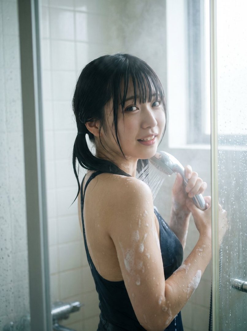 A young woman with black hair in twintails and bangs is showering, looking over her shoulder at the camera with a smile.