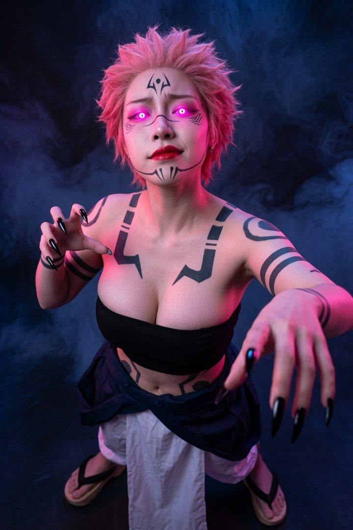 A woman cosplaying as Sukuna from Jujutsu Kaisen, with pink spiky hair, glowing purple eyes, and black body markings, posing in a studio with smoke.