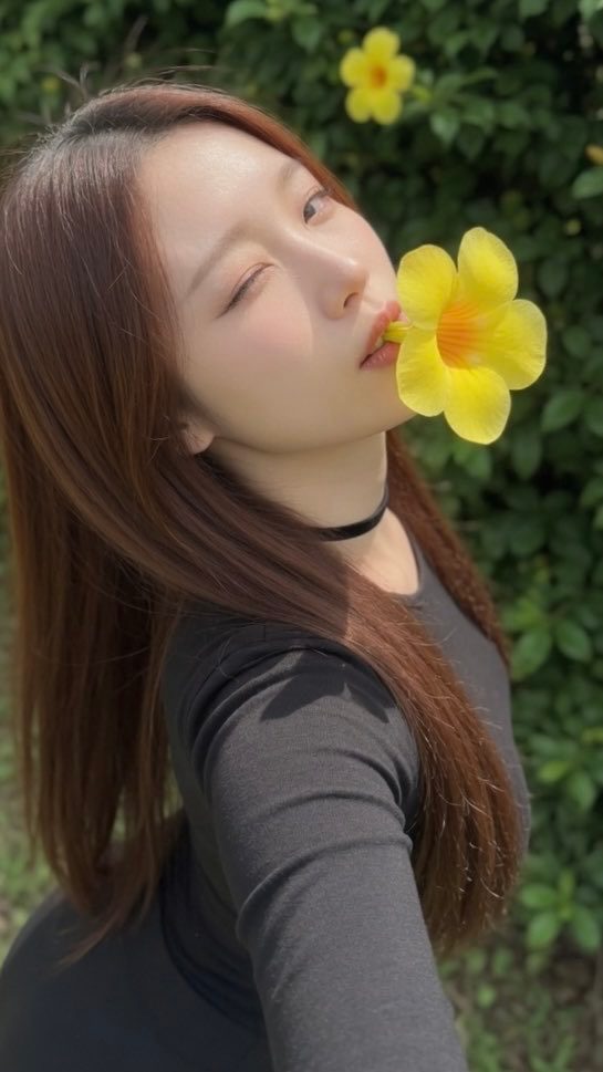 A young woman with long brown hair and a black top holds a yellow flower in her mouth, winking at the camera in an outdoor setting.