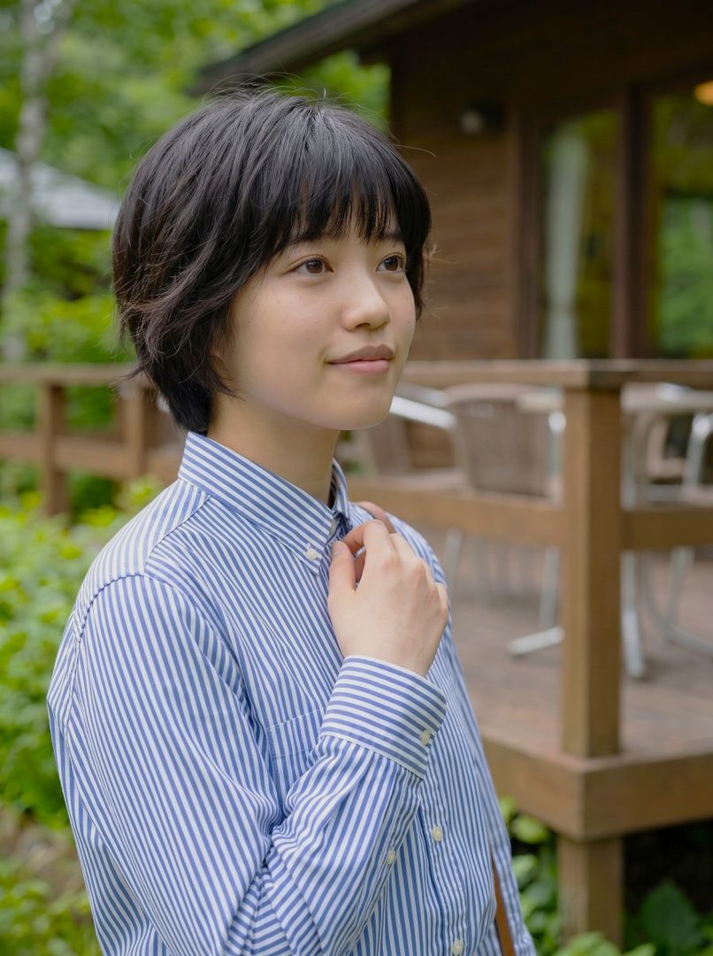 A young woman with short black hair and a striped blue and white shirt stands outdoors, looking to the side with a serene expression.
