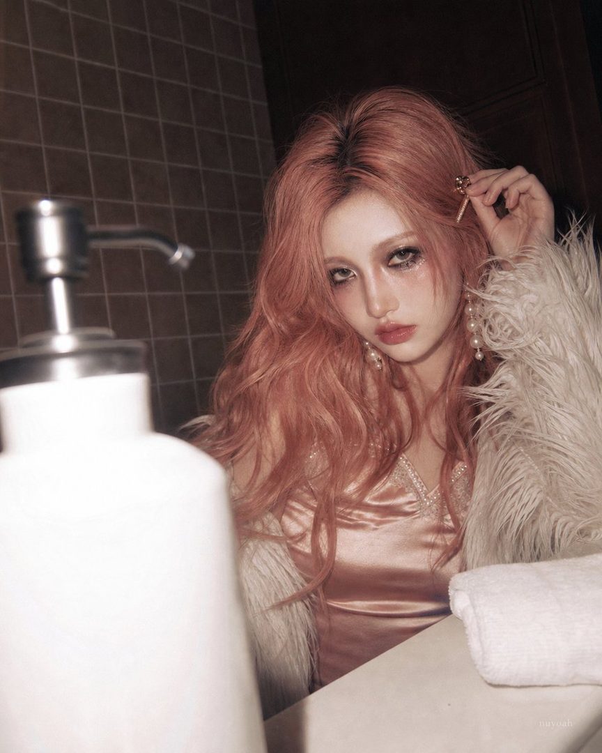 A woman with long pink hair and a satin dress looks directly at the camera in an indoor setting.