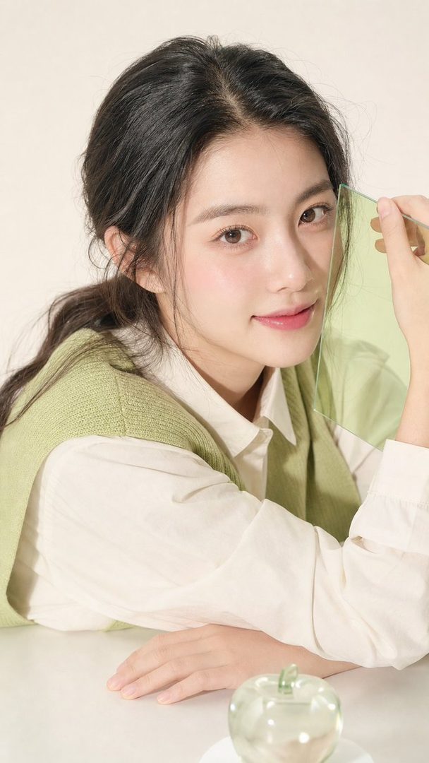A young woman with long black hair and a green knitted vest over a white shirt poses in a studio, holding a translucent green panel.