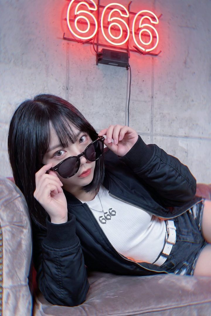 A young woman with black bob hair and bangs, wearing a black bomber jacket, white ribbed top with a '666' necklace, and denim shorts, poses on a couch with sunglasses pulled down, against a concrete wall with a red '666' neon sign.