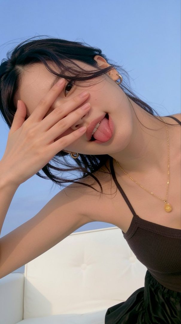 A playful East Asian woman with black hair covers her face with her hand and sticks out her tongue, wearing a dark top and a gold necklace against a clear blue sky.