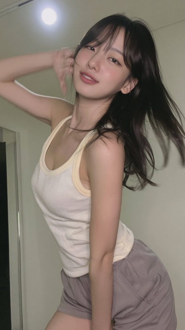 A young woman with long dark hair and bangs poses indoors, wearing a white tank top and grey shorts.