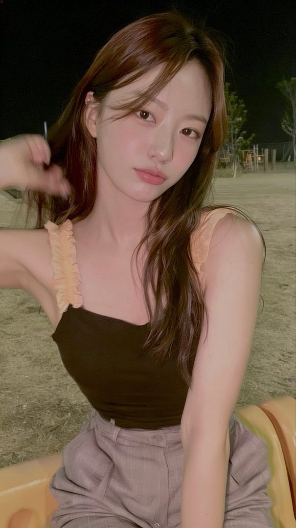 A photorealistic portrait of a young woman with long brown hair, wearing a dark top and light pants, sitting outdoors at night.