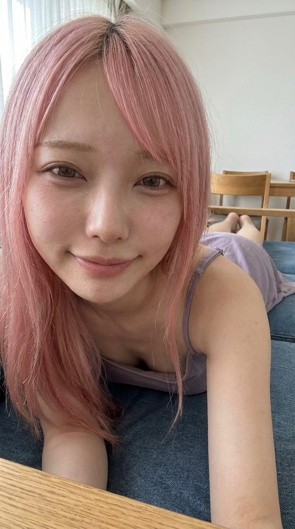 A woman with pink hair and bangs smiles gently while lying on her stomach, wearing a light purple camisole.