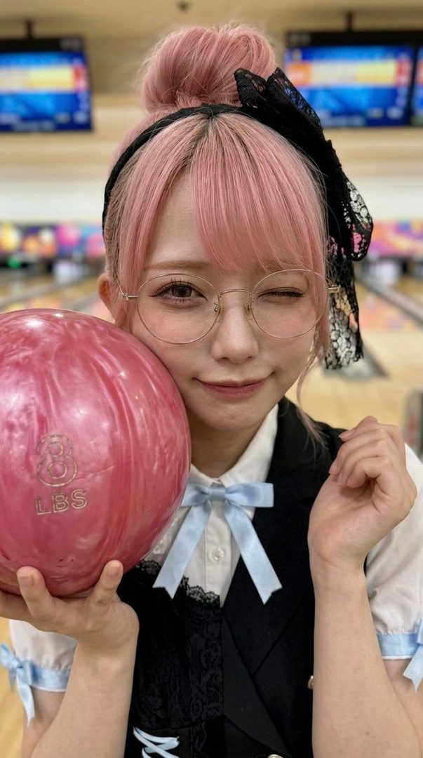 A young woman with pink hair in a bun and glasses winks while holding a pink bowling ball at a bowling alley.
