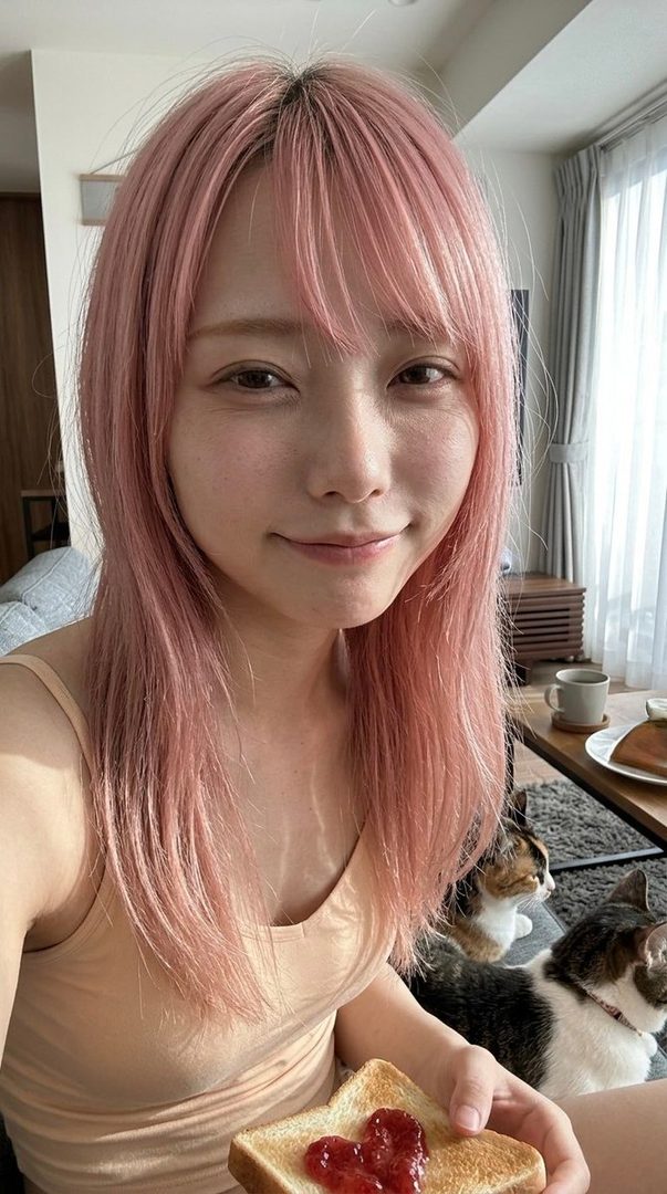 A young woman with pink hair and a tank top smiles while holding a piece of toast with heart-shaped jam, with two cats visible in the background.