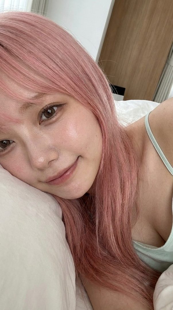 A close-up portrait of a woman with long pink hair and bangs, wearing a light blue camisole, lying on a white pillow with a gentle smile.