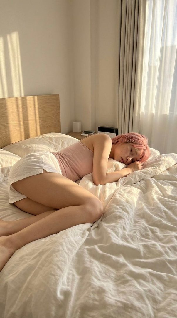 A person with pink hair is sleeping peacefully in bed, illuminated by soft morning light filtering through a window.
