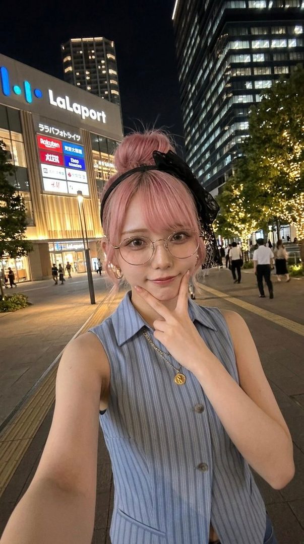 A young woman with pink hair and glasses poses with a peace sign on a city street at night, with a LaLaport building in the background.