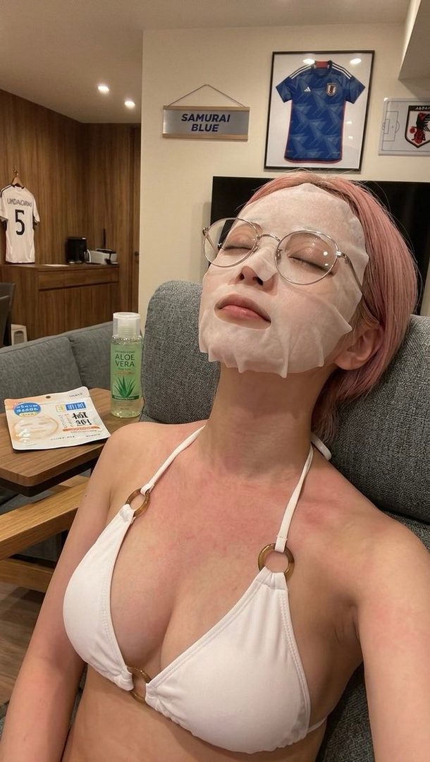 A person with pink hair and glasses is wearing a white bikini top and a face mask, relaxing indoors with a bottle of aloe vera and a soccer jersey visible in the background.