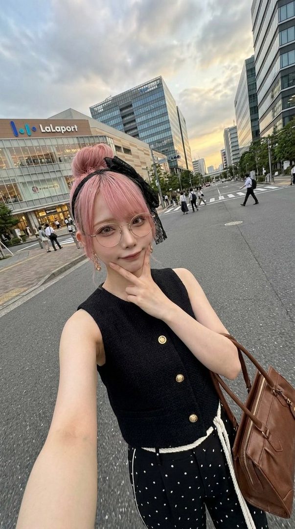 A young woman with pink hair in a bun and glasses takes a selfie on a city street with a shopping mall in the background.