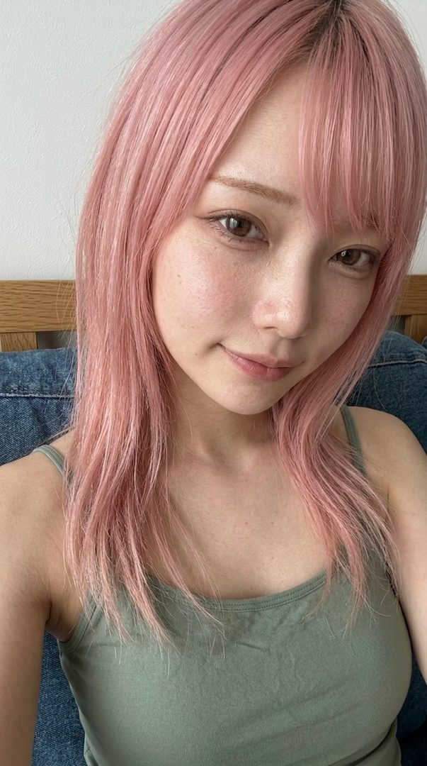 A woman with pink hair and bangs takes a selfie, wearing an olive green tank top indoors.