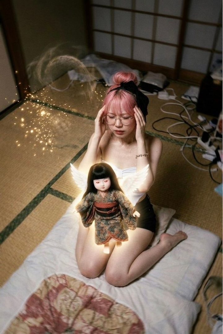 A woman with pink hair and glasses sits on a tatami mat, holding her head, with a glowing doll with wings on her lap, surrounded by sparkling effects.