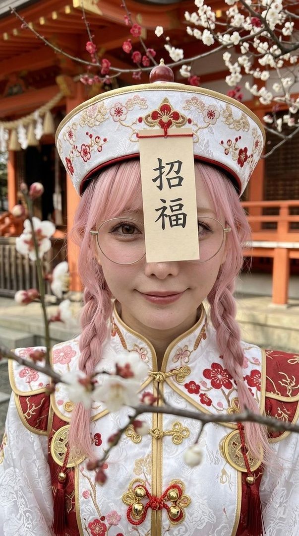 A young woman with pink braided hair and round glasses wears a white and red traditional Chinese dress and hat, standing in front of a Japanese temple with cherry blossoms.