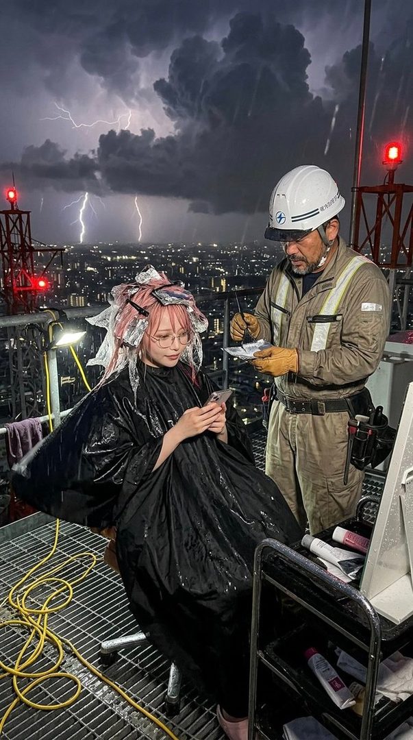 A person with pink hair and foils sits in a hairdressing cape, looking at their phone, while a stylist works on their hair on a rooftop during a lightning storm over a city at night.