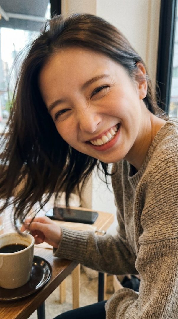 A young woman with dark brown hair smiles brightly while sitting at a cafe table with a cup of coffee.
