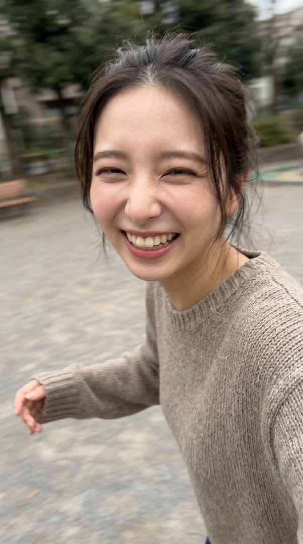 A young woman with dark brown hair tied in a messy bun, wearing a light brown knitted sweater, is captured mid-motion with a wide, joyful smile in an outdoor park setting, featuring a slight motion blur.