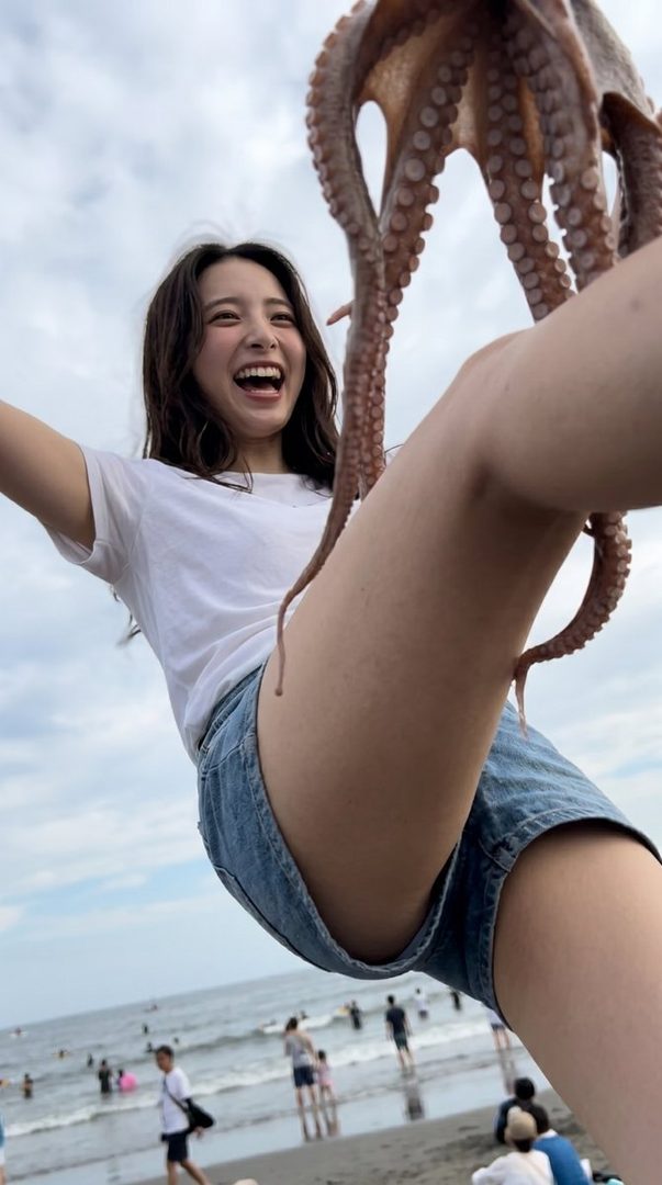 A young woman in a white t-shirt and denim shorts laughs joyfully while holding an octopus at a crowded beach.