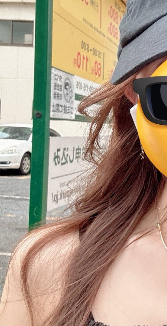 A close-up selfie of a person with long brown hair, wearing a hat and sunglasses, with a street scene in the background.