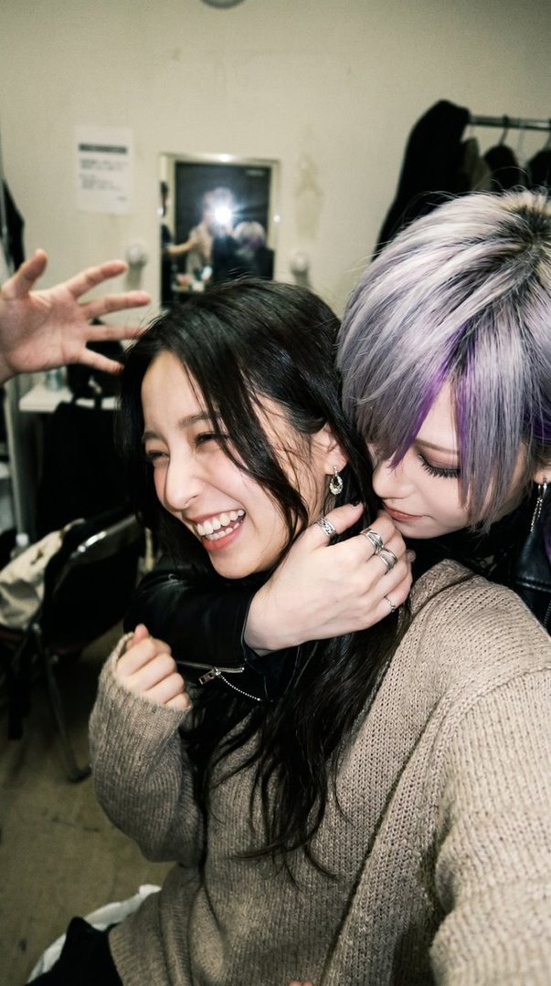 Two women are playfully embracing, one with long dark hair laughing and the other with short silver hair leaning in affectionately, in what appears to be a backstage or dressing room setting.
