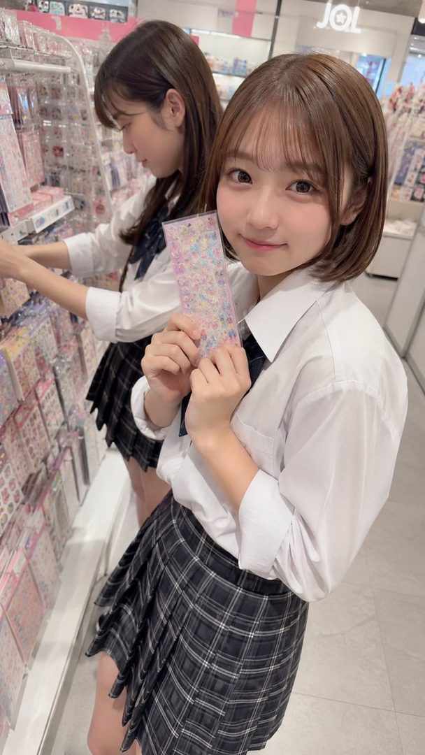 Two young women in school uniforms are shopping for stickers in a store, with one holding up a pack of stickers and smiling at the camera.