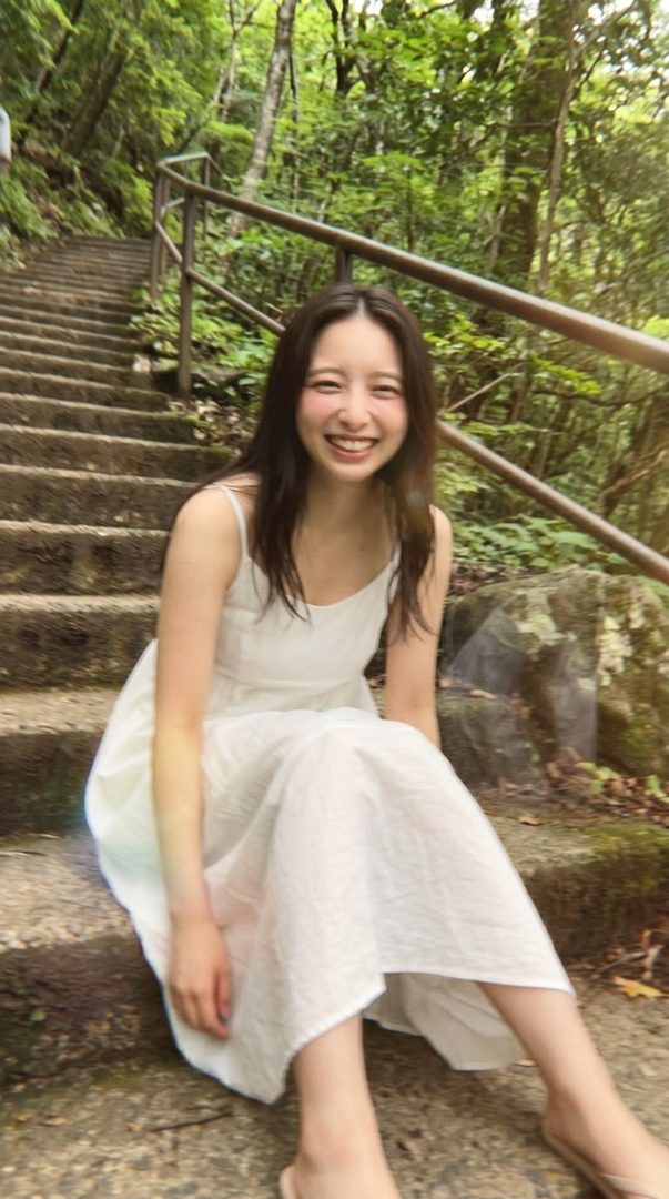 A smiling woman with long dark hair sits on stone steps in a forest, wearing a white sundress.