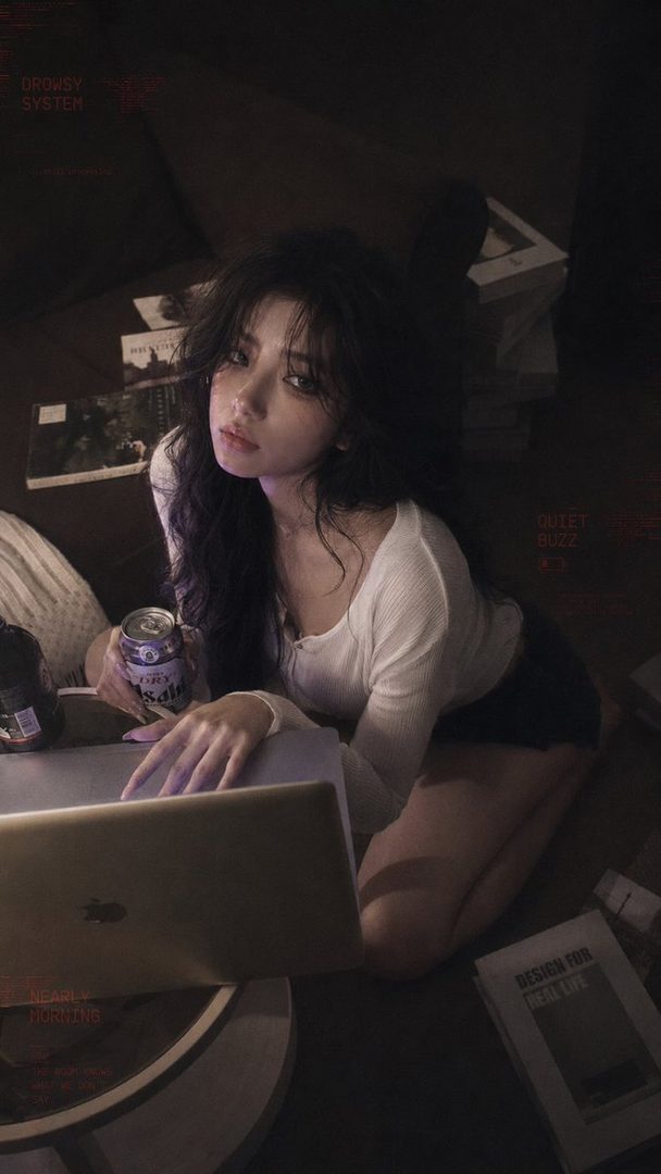 A young woman with long dark hair is kneeling on the floor, looking at the camera while holding a can and resting her hand on a laptop.