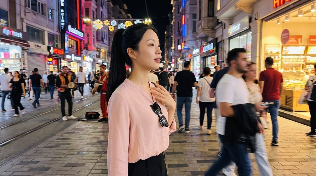 A young woman with black hair in a ponytail stands on a bustling city street at night, wearing a pink blouse and black pants.