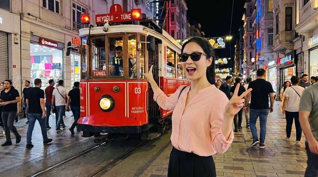 A young woman in sunglasses makes a peace sign in front of a red tram on a bustling city street at night.