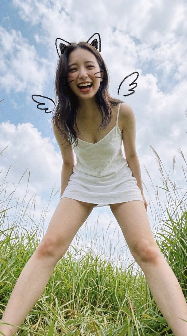 A young woman with dark brown hair, wearing a white slip dress and a cat ear headband, crouches in a grassy field under a blue sky, laughing playfully.